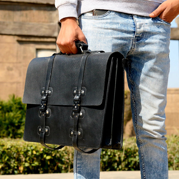 A person holding a black leather briefcase with brass hardware and a zipper closure. The bag appears to have a sleek and professional design.