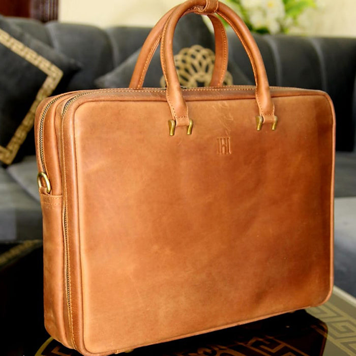 A brown leather laptop briefcase with a slim design, featuring a locking mechanism, two handles, and a detachable shoulder strap.