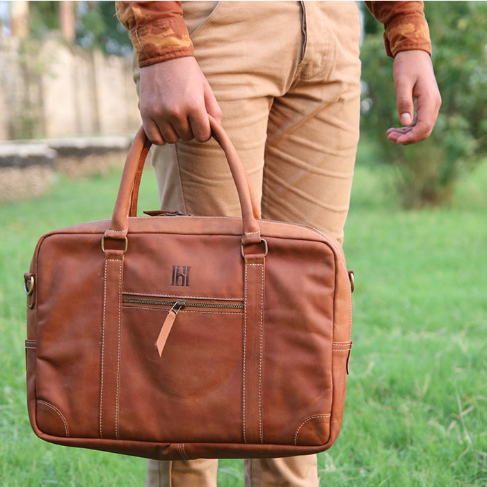 A person holding a brown leather laptop bag with brass hardware, featuring a sleek and professional design with quick-access pockets.